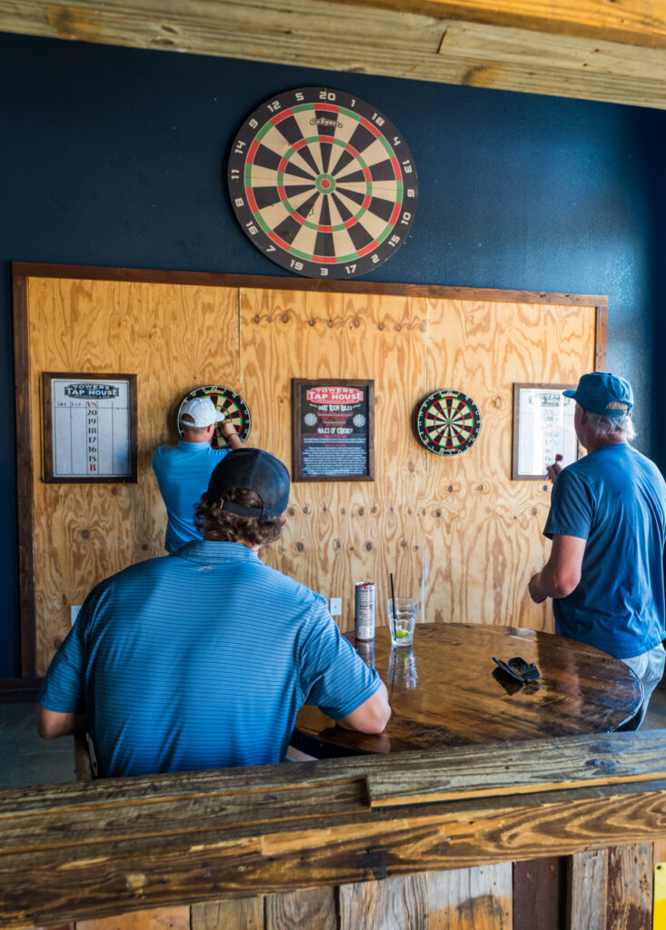 Three men play darts in a wood-paneled bar, with several dartboards and scoreboards on the wall and a large board above them.