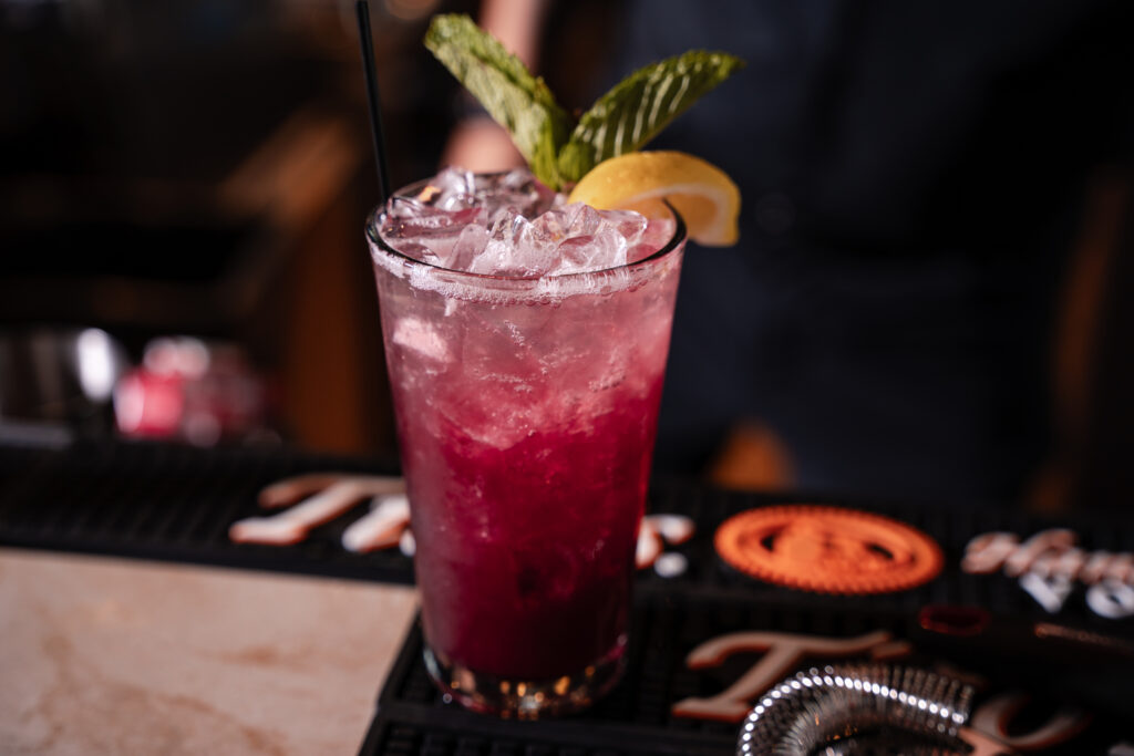 A close-up view of a vibrant purple cocktail in a tall glass, garnished with a lemon wedge, fresh mint sprigs, and a black straw, resting on a bar mat.