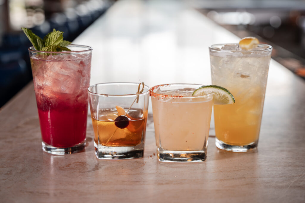 Four colorful cocktails in various glasses sit in a line on a light-colored bar top, each garnished differently with mint, a cherry, a lime wedge, or a lemon wedge.