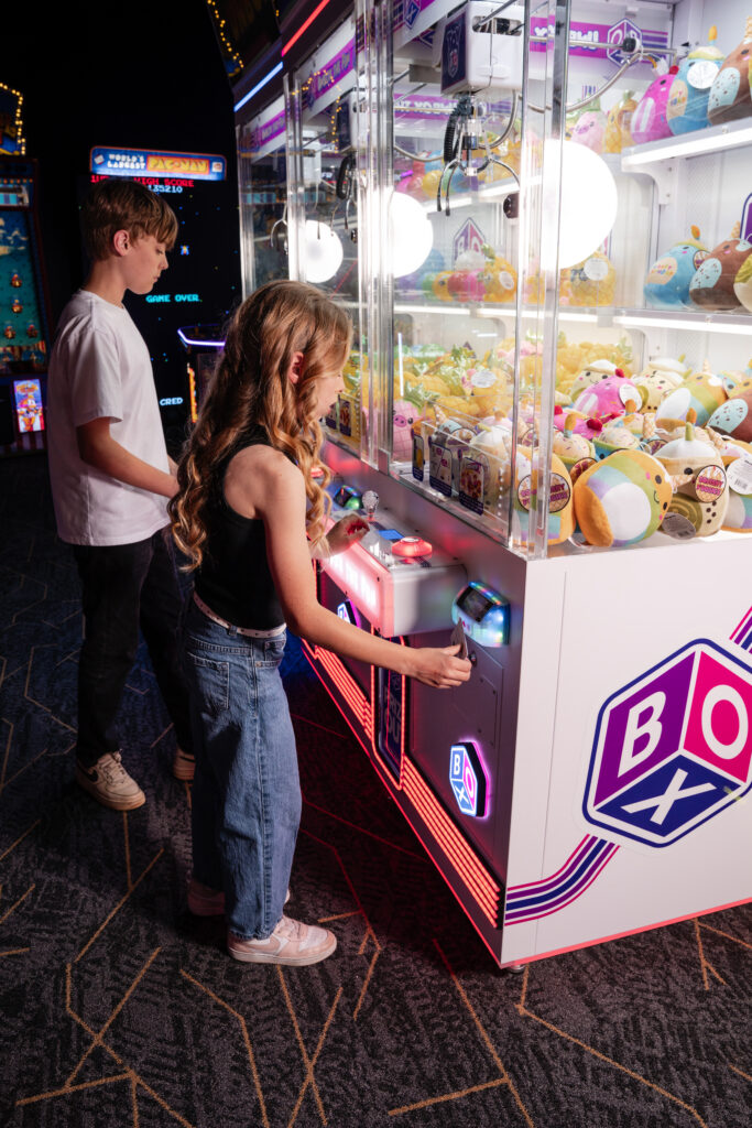 Two children stand at a brightly lit claw machine inside an arcade, with the girl in the foreground inserting a coin or card to play while stuffed animals fill the display case.