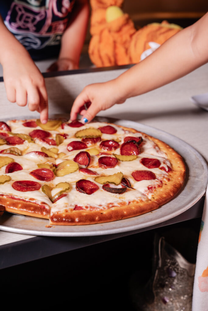 Two children stand over a round silver tray holding a cooked pizza topped with melted cheese, pepperoni slices, and pickle slices, each child reaching down with a hand to adjust a pickle.