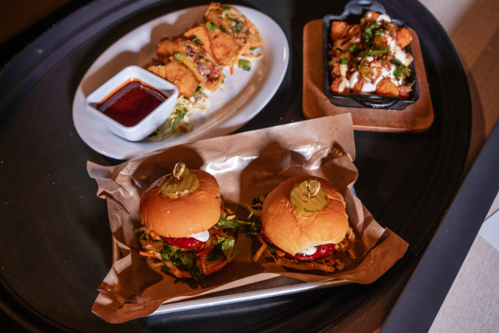 An overhead shot of a dark serving tray holding a plate of fried spring rolls with a dipping sauce, a cast-iron skillet of loaded tater tots, and two sliders served on brown paper with pickles on top.