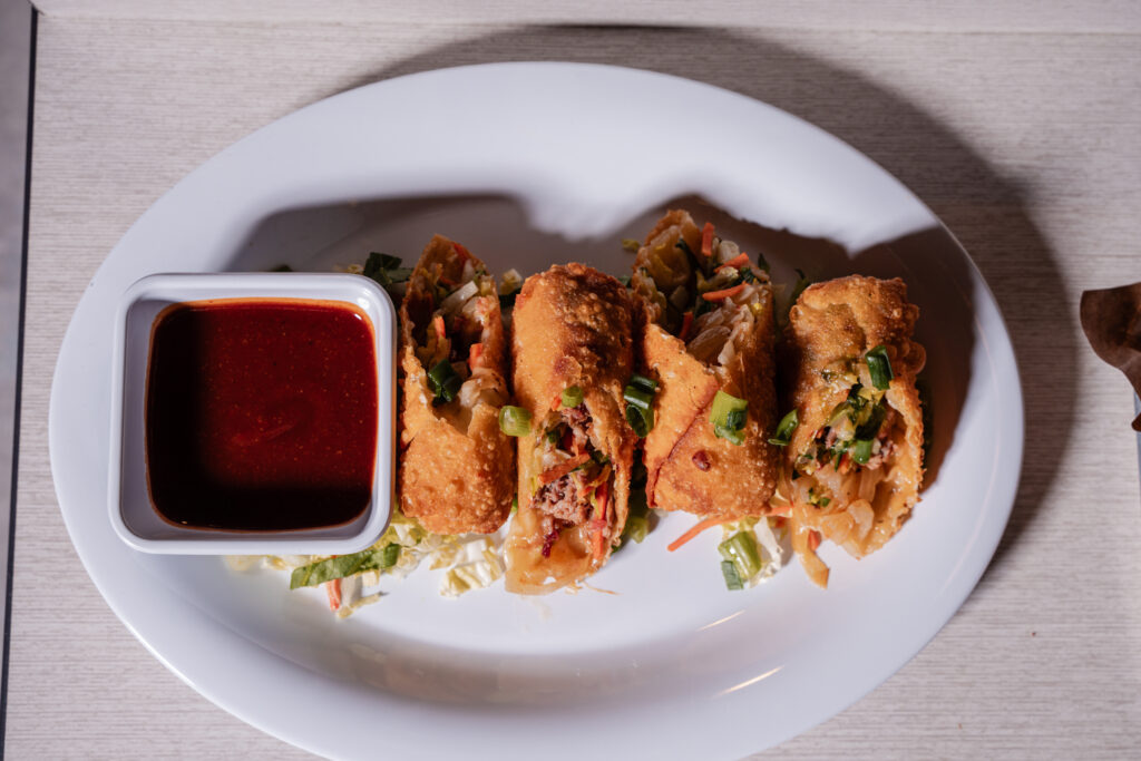 An overhead shot of four golden-brown fried spring rolls on a bed of shredded cabbage on a white oval plate, accompanied by a small square bowl of red dipping sauce.