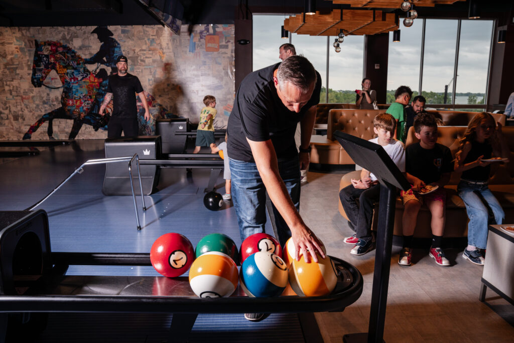 A man in a black shirt and jeans reaches for a bowling ball on a rack inside an upscale bowling alley, while several children and adults watch from nearby seating against a backdrop of large windows and a large mural of a horse.