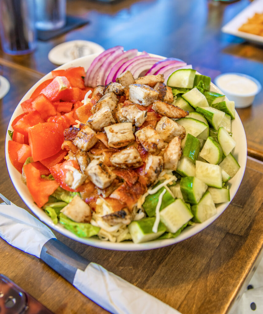 Bowl of salad topped with grilled chicken, tomatoes, cucumbers, red onion, lettuce, and pita pieces.