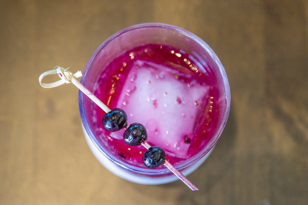 Top-down view of a bright magenta cocktail with ice and a skewer holding three dark berries on a wooden table