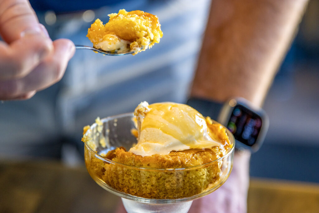 Close-up of a spoon lifting caramel-topped ice cream and cake from a glass dessert dish.