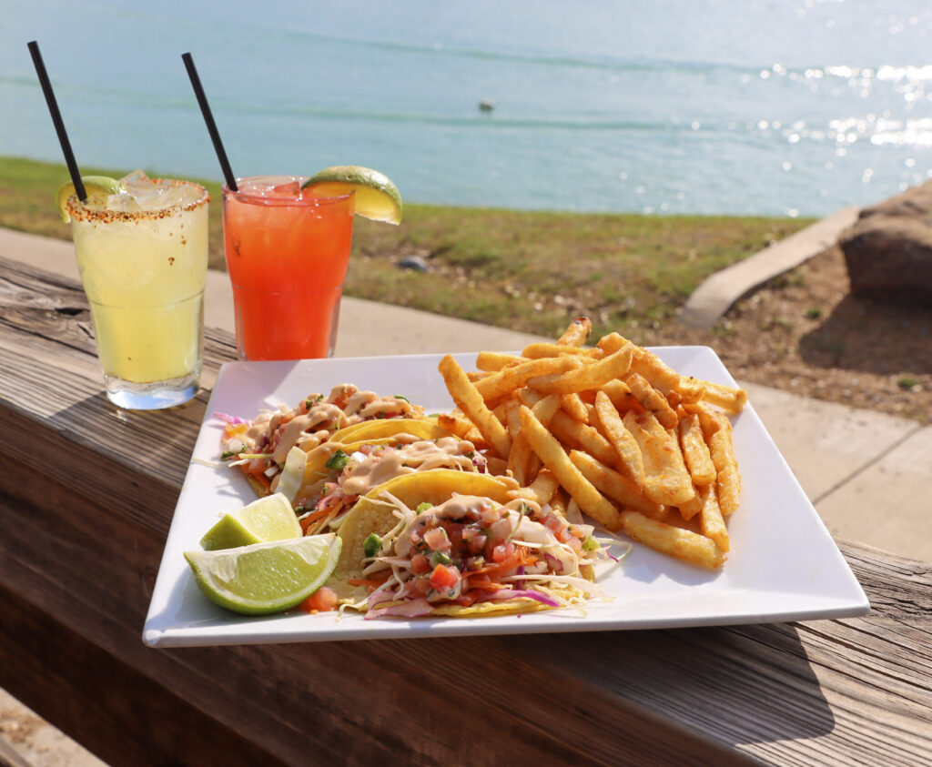 A plate of fish tacos, fries, and lime wedges sits on a wooden seaside railing beside two colorful drinks, with the ocean behind.