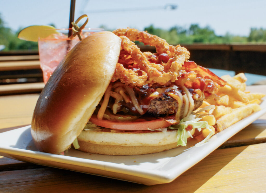 A bacon burger topped with crispy onion rings and served with French fries sits on a white plate on a wooden picnic table.