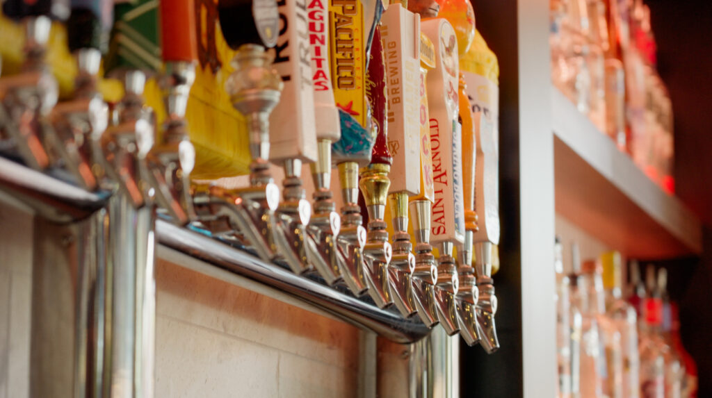 A row of colorful beer tap handles and metal spouts lines a bar, with bottles and shelves visible in the background.