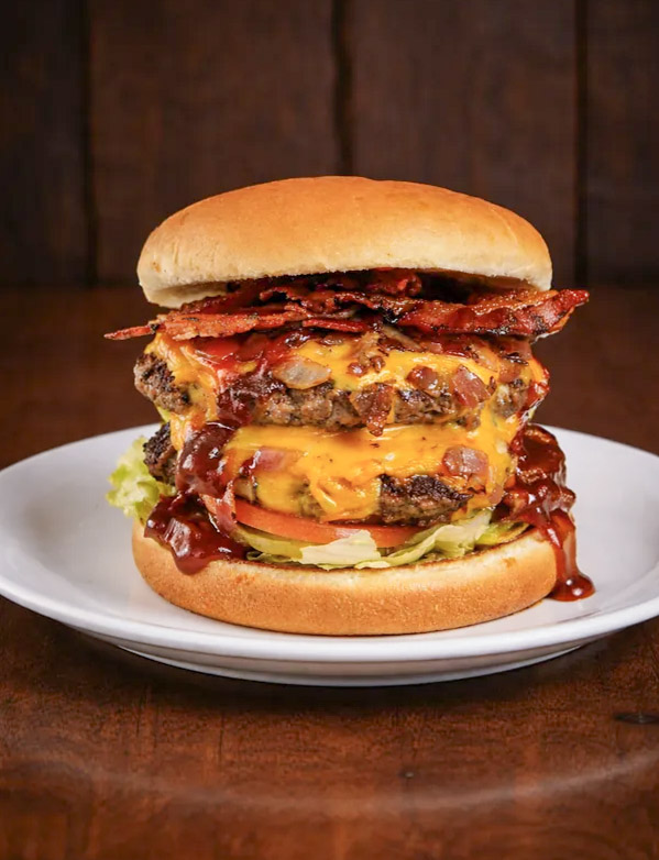 A double cheeseburger with bacon, lettuce, and tomato sits on a white plate against a dark wooden background.