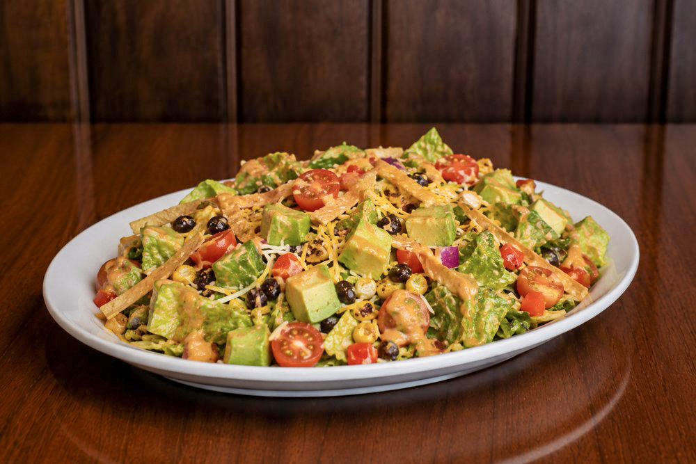 A white oval plate of taco salad with lettuce, avocado, cherry tomatoes, black beans, and shredded cheese on a wooden table.