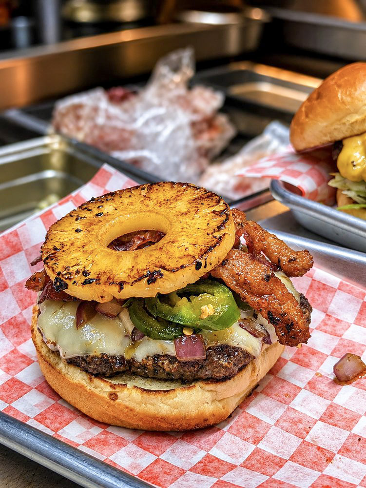 A cheeseburger topped with a grilled pineapple ring and bacon sits on red-and-white check paper in a metal tray, with another burger blurred in the background.