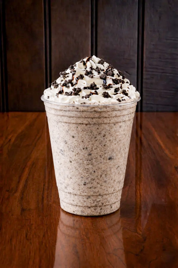 A cookies and cream milkshake topped with whipped cream and chocolate pieces sits in a clear plastic cup on a wooden table.